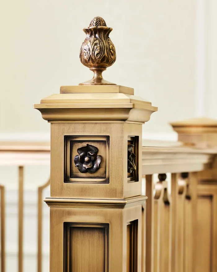 A finished architectural bronze newel post with a cast flower relief panel set into the post and an ornamental finial above, the balusters running away behind it