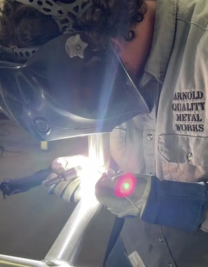 A welder under the hood, TIG arc lit, running a bead on an aluminum frame in the shop