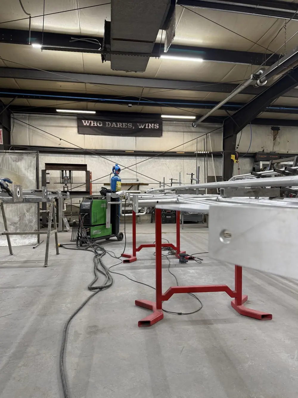 The shop floor with aluminum assemblies on stands, a banner reading WHO DARES, WINS on the far wall