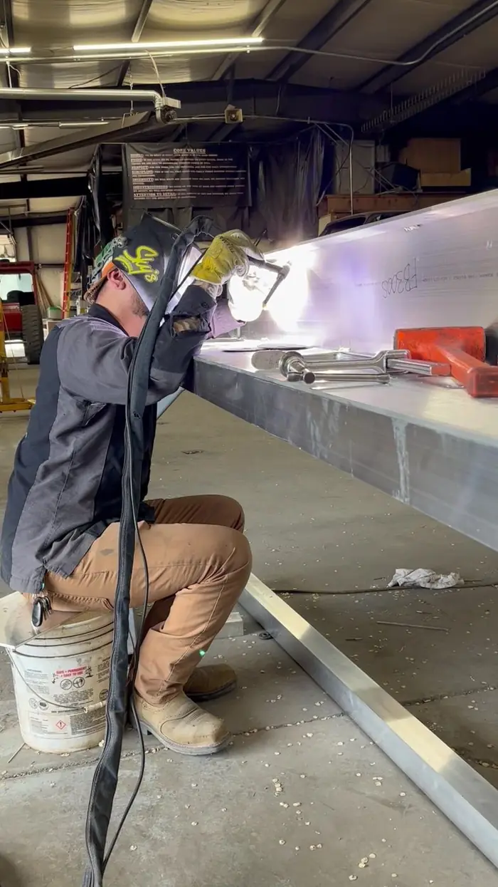 A welder kneeling to run a long seam down an aluminum assembly