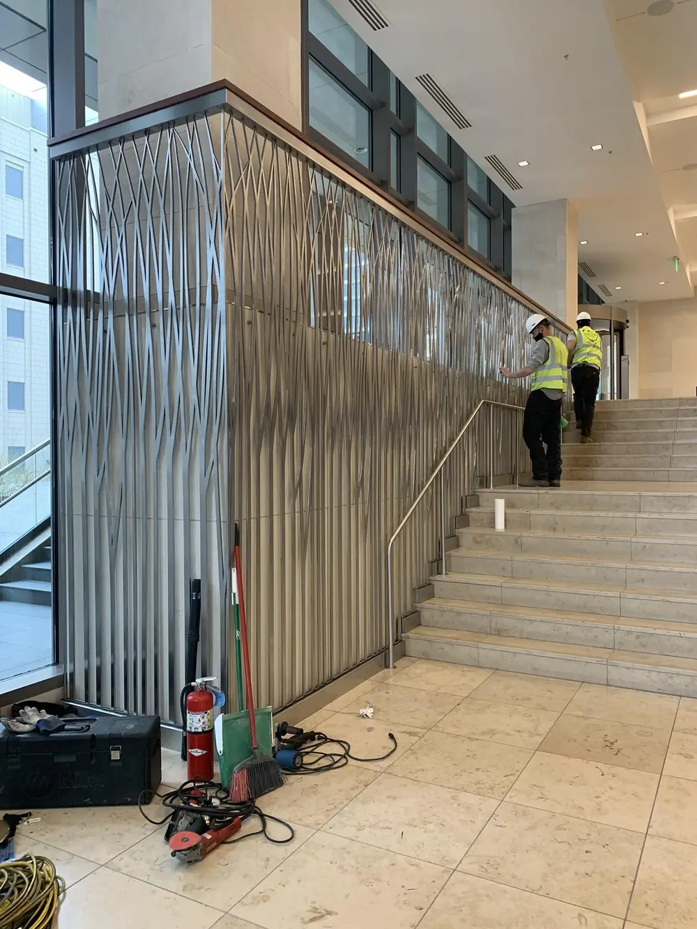 Full-height stainless grass-motif panel rising alongside the stair during installation, crew working at the top