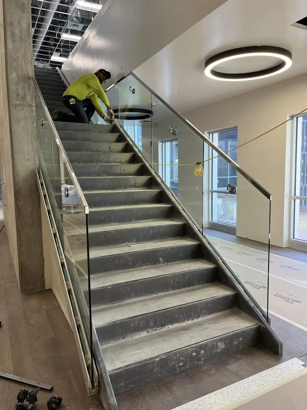 A welder kneeling on a stair setting a glass guardrail panel into its base shoe