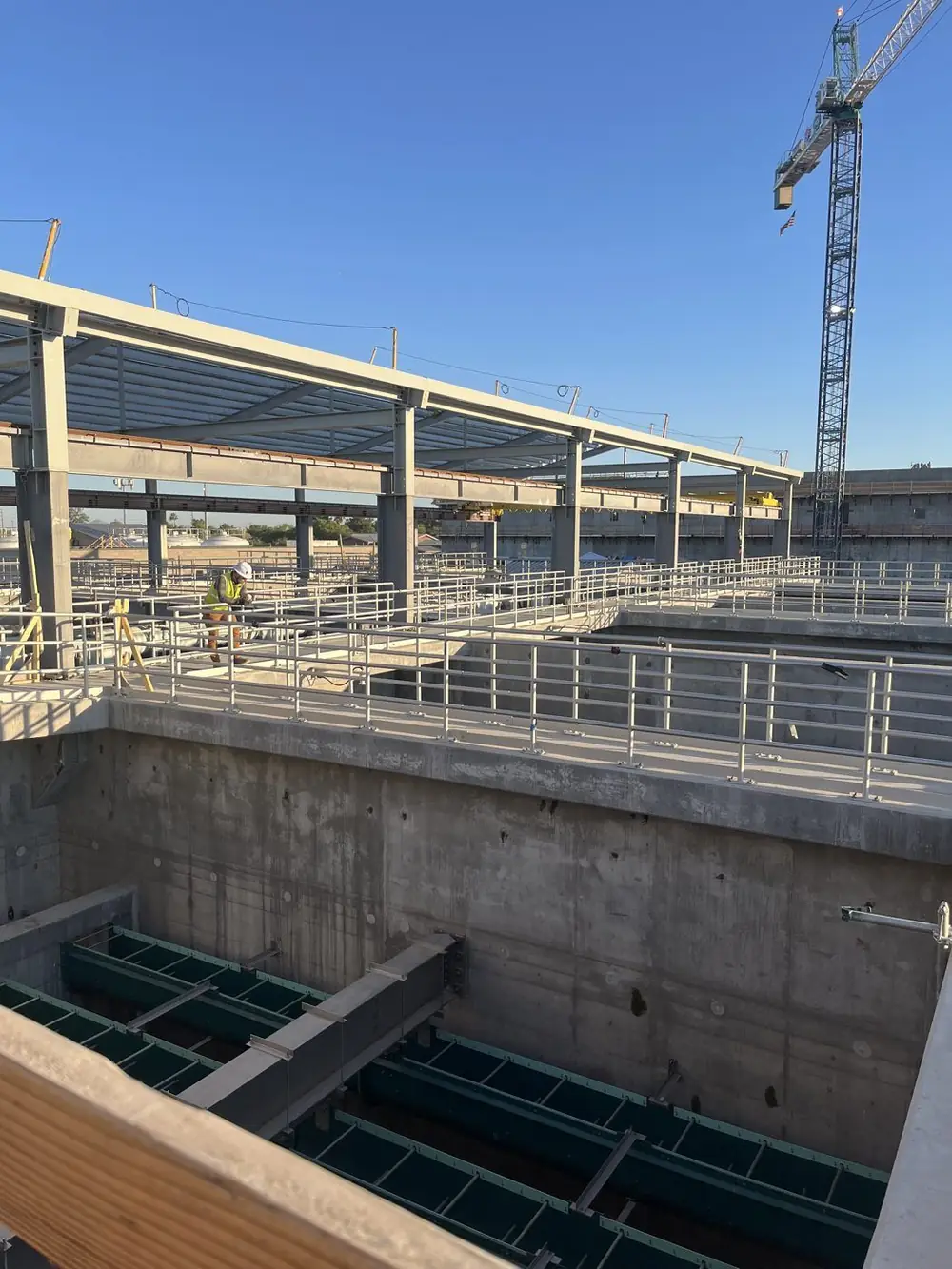 Wide view of the Gilbert plant under construction — long runs of anodized aluminum guardrail along concrete basins beneath a tower crane