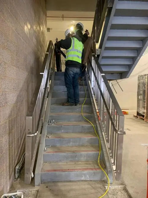 Two Arnold Quality crew members installing stainless stair handrail on site in hard hats and high-visibility vests